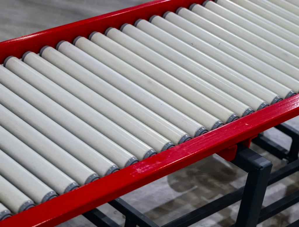 Red-framed roller conveyor with white cylindrical rollers used for material handling in an industrial setting.