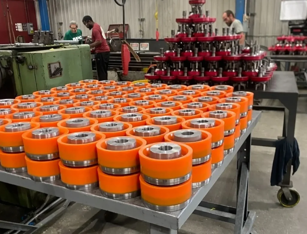 Rows of orange urethane rollers arranged on a metal table inside a manufacturing facility with workers and machinery in the background.